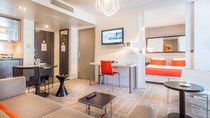 Una habitación de hotel moderna con comedor, escritorio, cama con ropa de cama naranja y televisor montado en la pared.