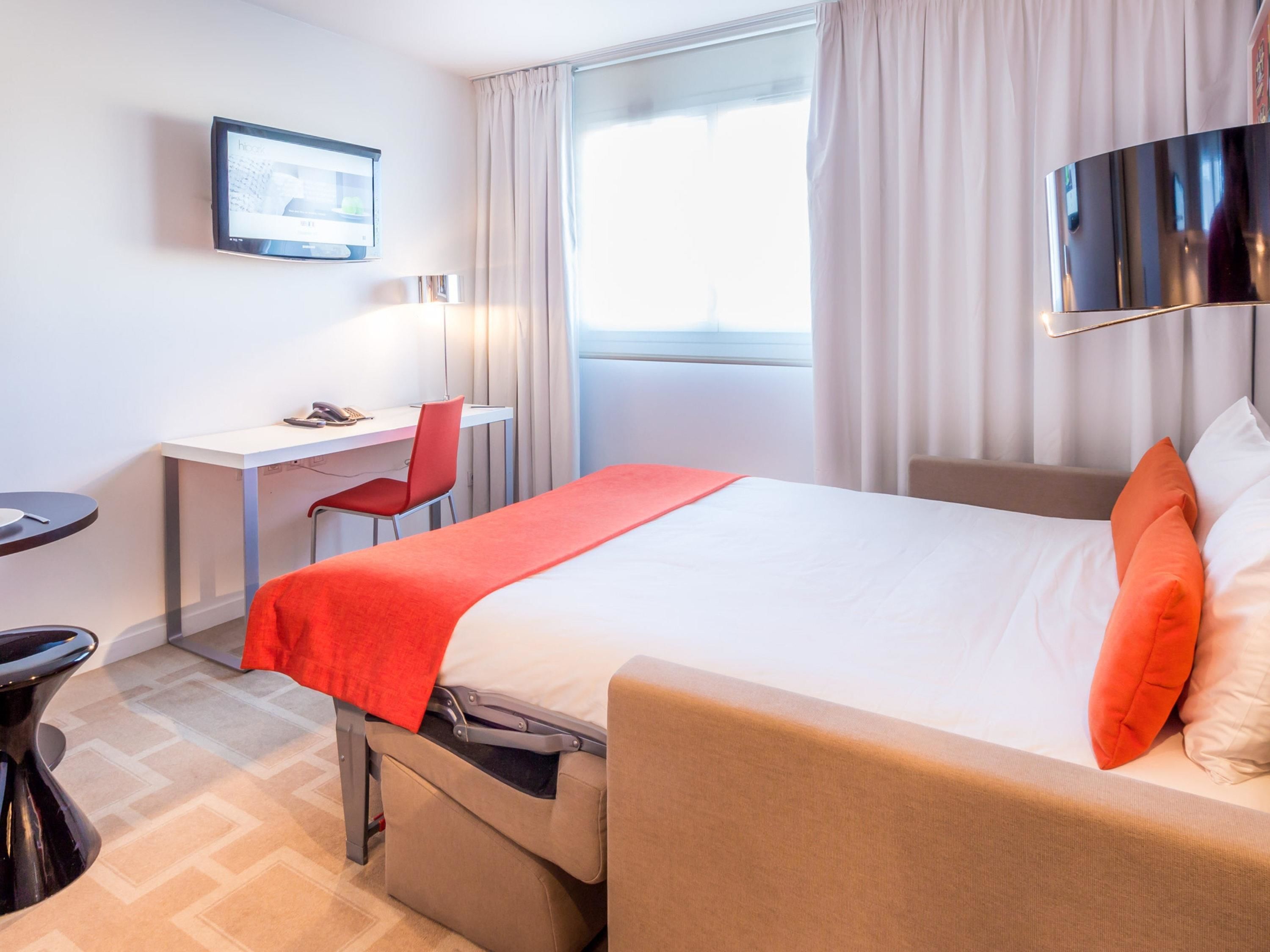 A hotel room with a bed, a desk, a chair, a TV, and a window with curtains.