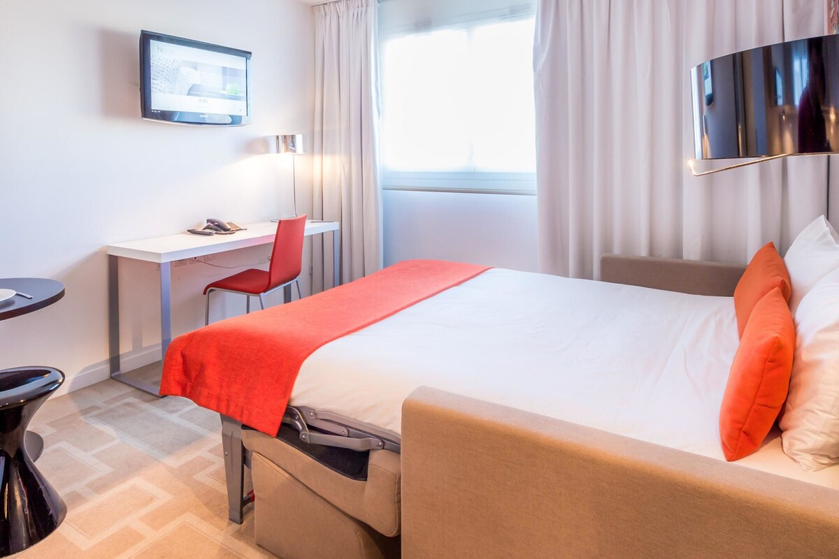 Habitación de hotel con cama, escritorio, silla, televisor y ventana con cortinas.