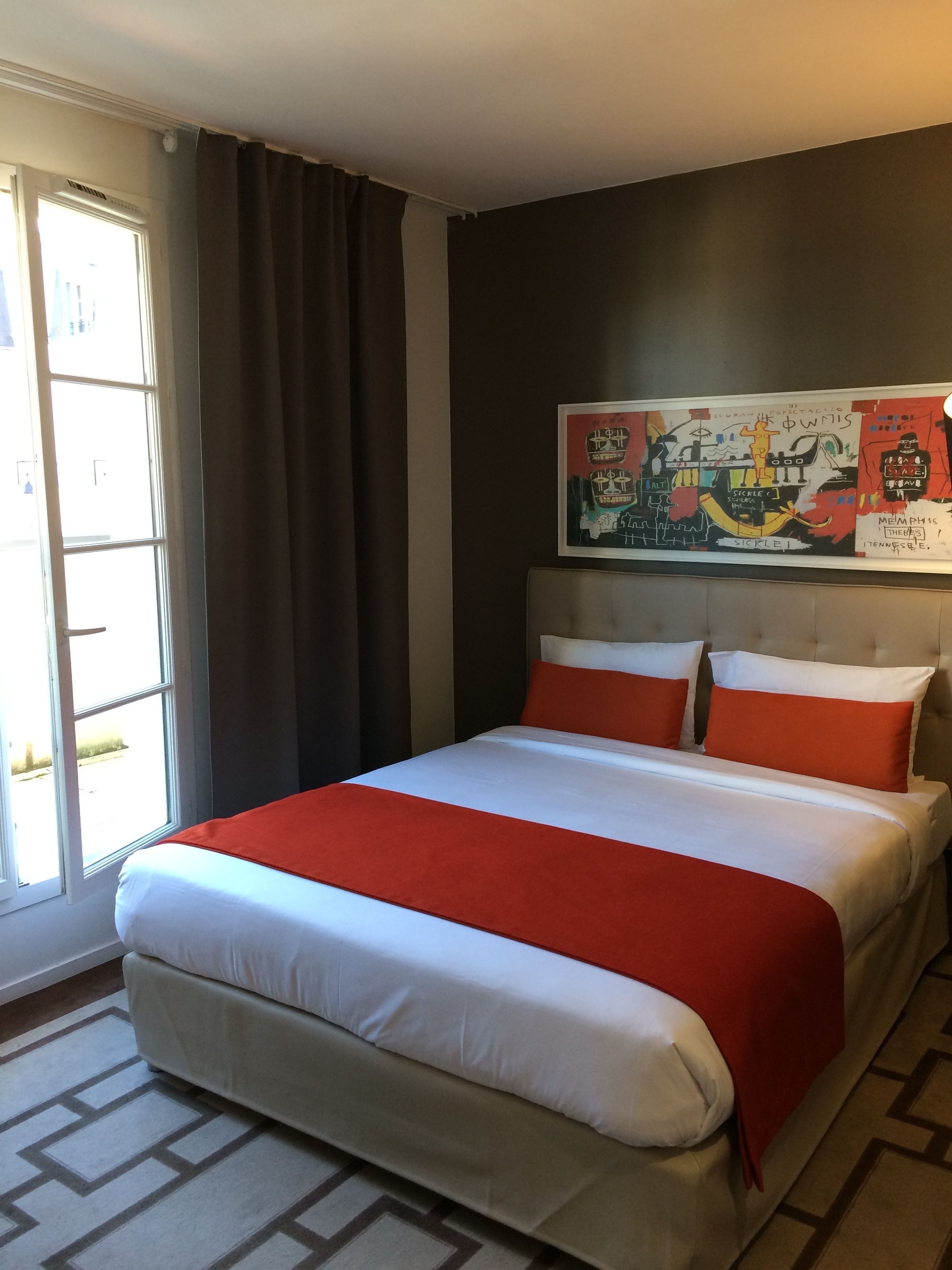 A hotel room with a large bed, a headboard with artwork, a window with curtains, and a patterned rug.