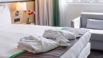 A hotel room with a neatly made bed, a bedside lamp, a vase with flowers, and a view of a cityscape through the window.