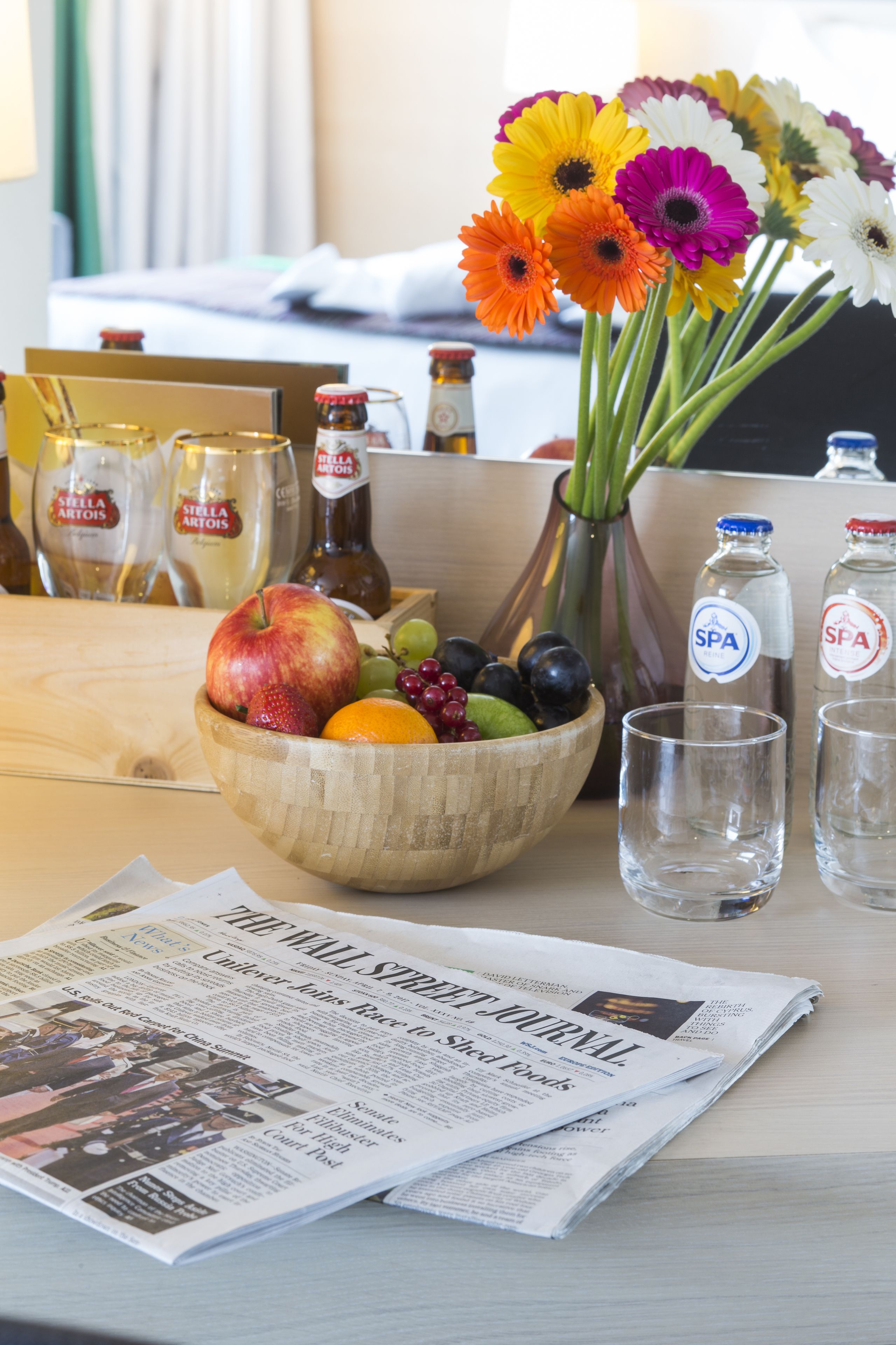 A wooden bowl with fruits, a vase with flowers, and two bottles labeled SPA on a table with a newspaper.