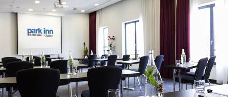 Un salón de conferencias con una pantalla de proyector mostrando el logotipo de 'park inn', sillas negras, mesas con botellas de agua y un vaso de agua sobre la mesa.