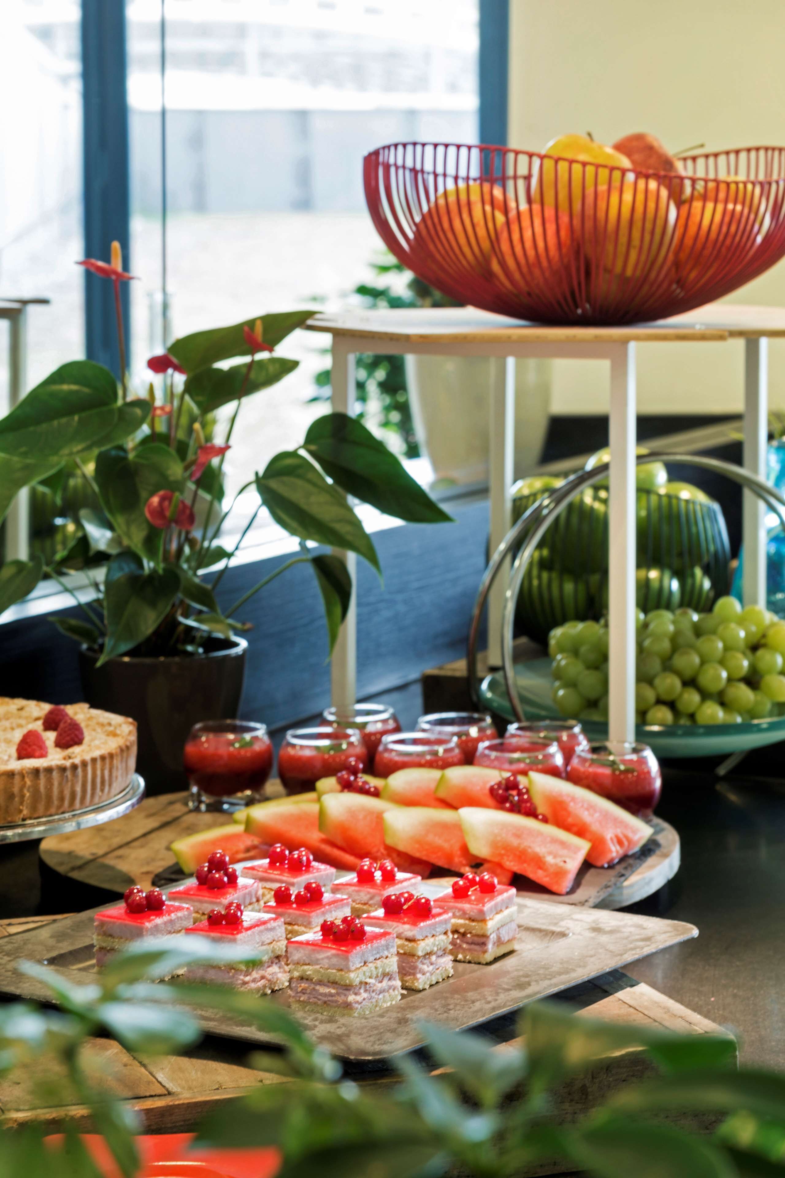 Una exhibición de frutas y postres sobre una mesa de madera, con una planta en maceta y un bol de naranjas al fondo.