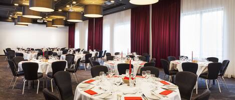 Un salón de banquetes con mesas redondas dispuestas para comer, sillas negras y un techo moderno con lámparas colgantes.
