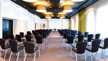 A conference room with rows of chairs facing a presentation screen, a projector, and a large window with curtains.
