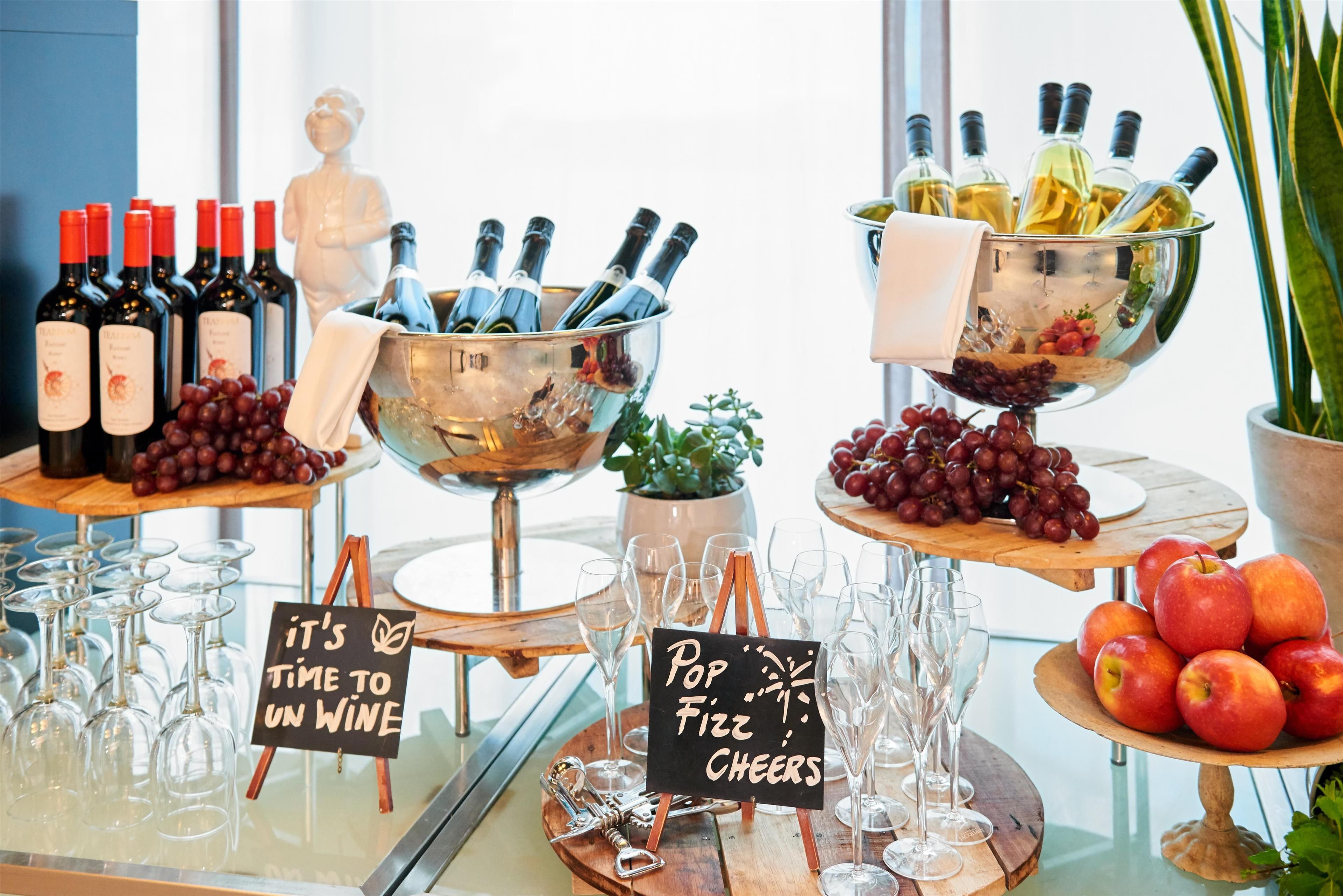 A bar area with wine bottles, a champagne bucket, and a bowl of grapes. There are also empty wine glasses and a sign that says 'Pop Fizz Cheers'.