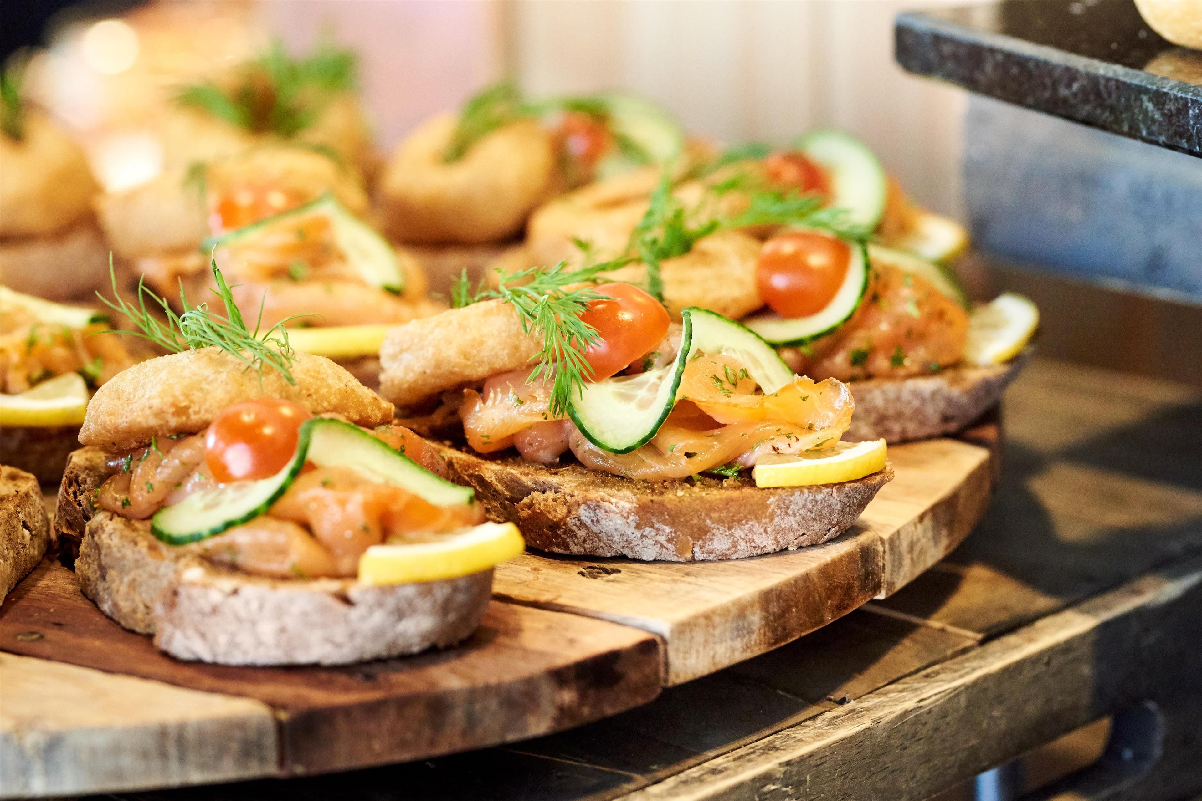 A wooden board with several open-faced sandwiches topped with fish, tomatoes, cucumbers, and herbs.