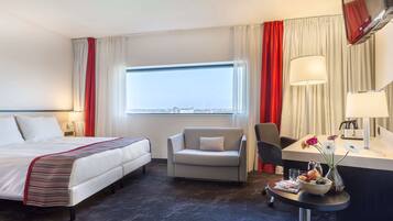 Una habitación de hotel con cama, escritorio con silla, una mesita con un florero y una ventana con vista a la ciudad.