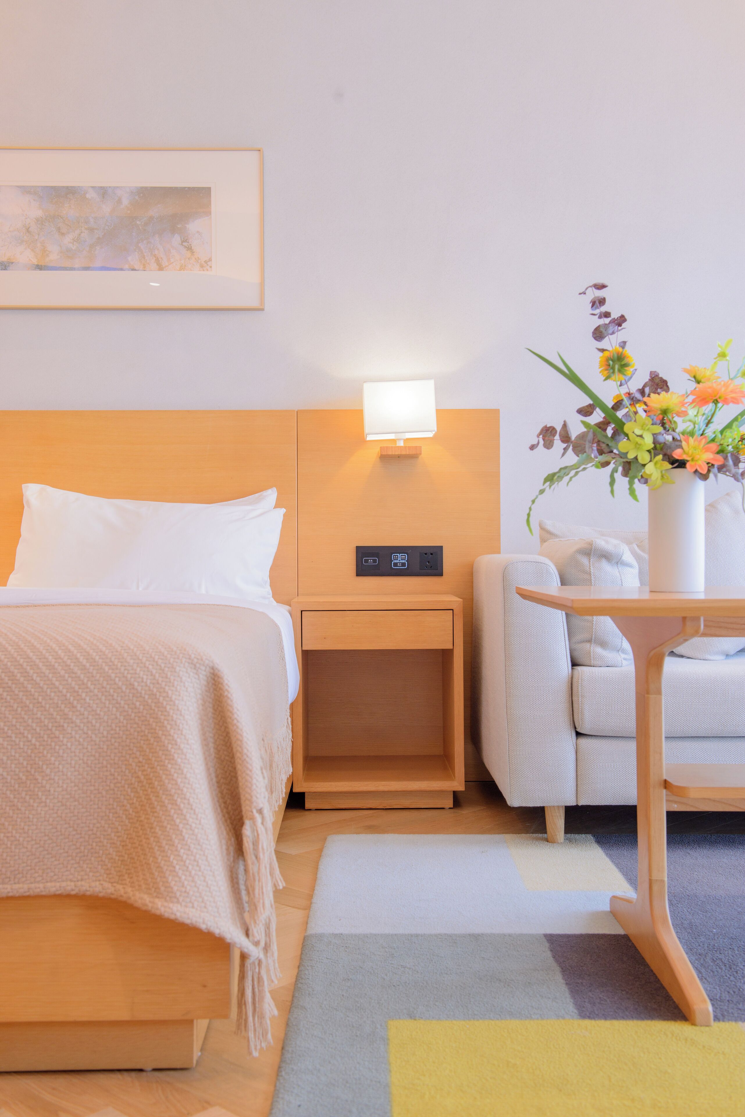 Et hotelværelse med en seng, et sengebord, en lampe, en stol og en vase med blomster.