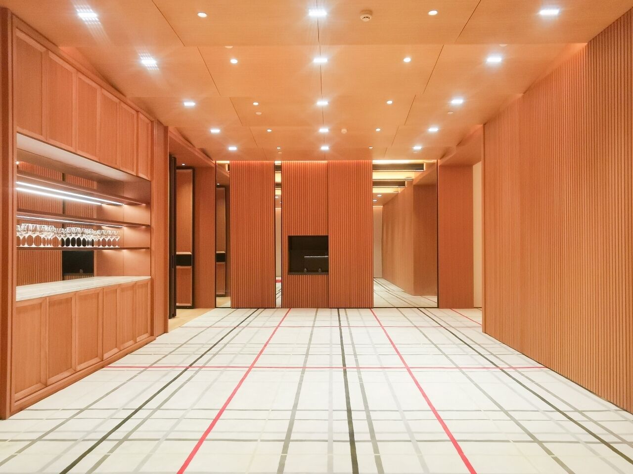 A corridor with wooden panel walls, recessed lighting, and a tiled floor with red lines.