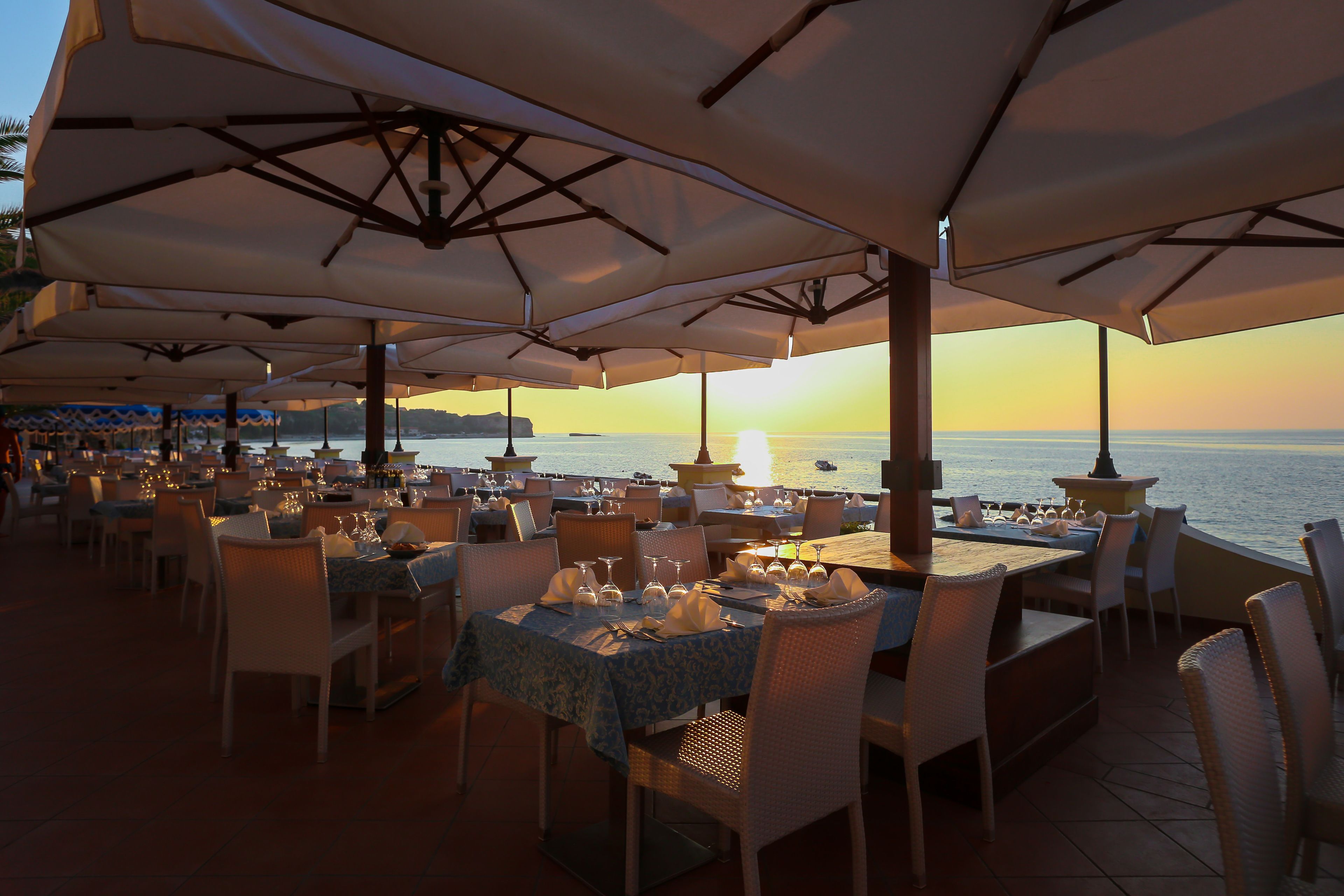 Un espacio al aire libre para comer con mesas y sillas, sombrillas y vistas al mar al atardecer.