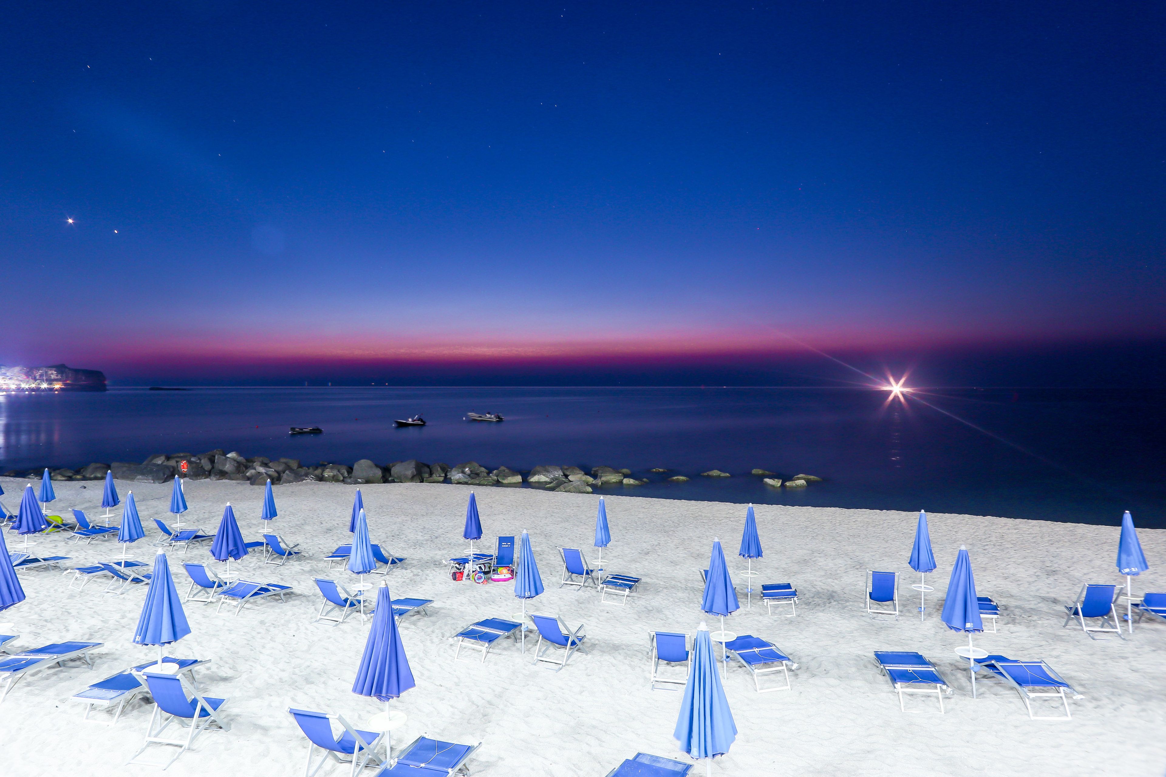 Una spiaggia con file di sdraio azzurre e ombrelloni, un mare calmo e un cielo al tramonto.