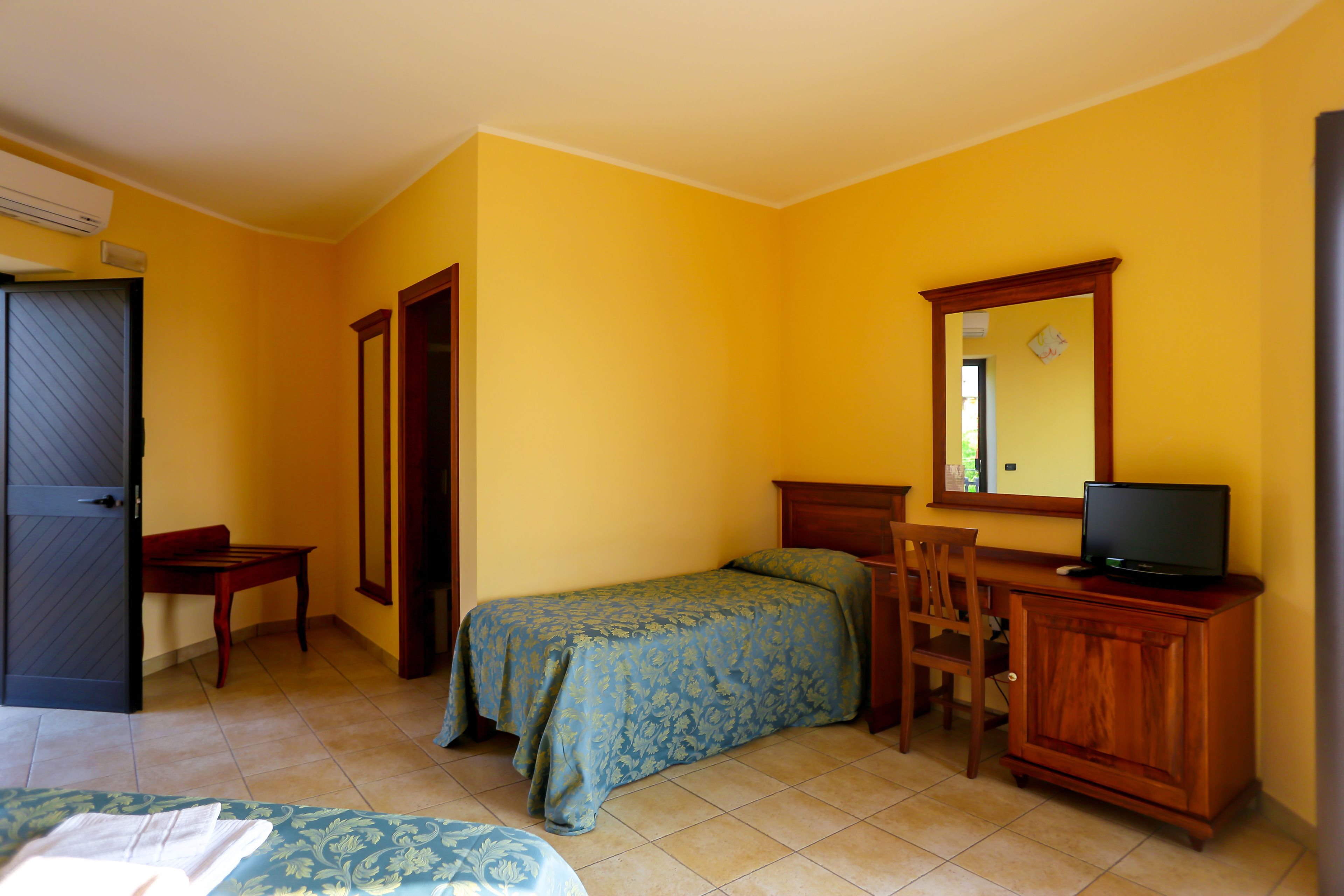A room with a bed, a desk, a chair, a television on a wooden cabinet, and a mirror on the wall.