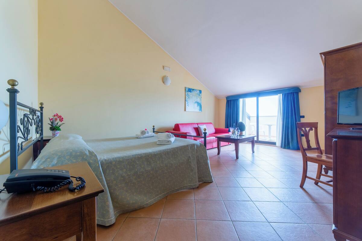 Una camera d'albergo con un letto, una scrivania con telefono, un divano rosso, una sedia di legno e un televisore.