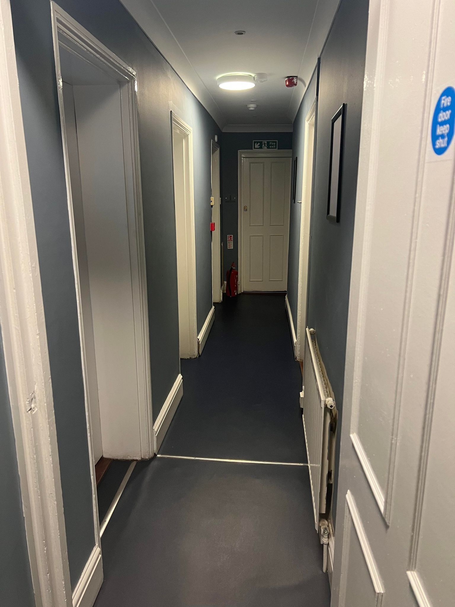 Un couloir étroit, avec des portes de chaque côté, un panneau indiquant la sortie de secours et un extincteur.