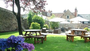 Une terrasse de jardin avec des tables et des bancs en bois, entourée d’un mur de pierre et d’une pelouse verdoyante.