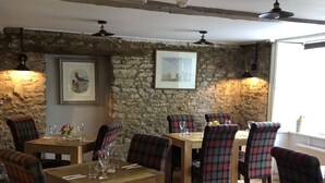 Un restaurant avec des tables en bois et des chaises à carreaux, des murs en pierre et des œuvres d’art encadrées.