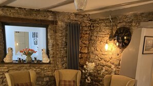 Un salon chaleureux avec des murs en pierre, une cheminée et une grande fenêtre. On y trouve trois fauteuils et une table basse ronde sur laquelle est posé un saladier.