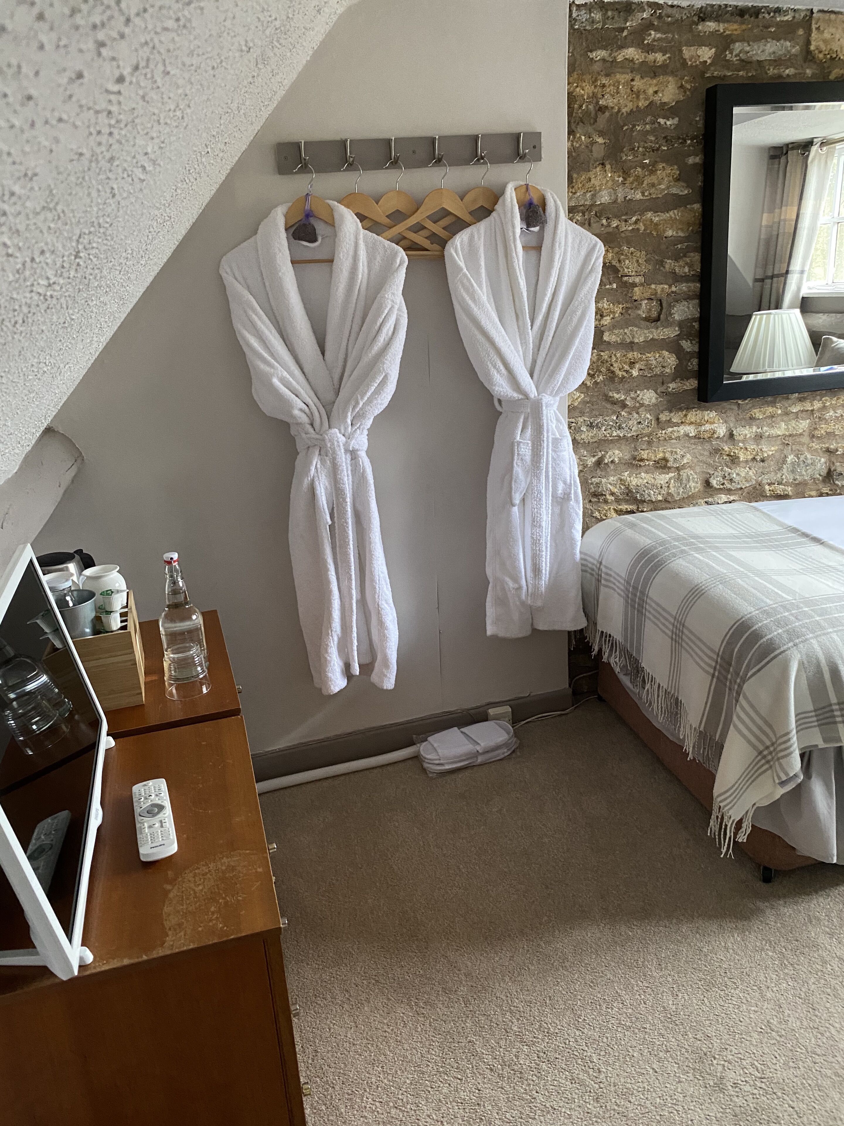 Quarto de hotel com cama, penteadeira de madeira, escrivaninha com um laptop e controles remotos, e dois roupões brancos pendurados em um suporte.