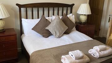 Una cama bien hecha con cabecera marrón, ropa de cama blanca y varios almohadones. Hay dos mesitas de noche con lámparas, un atril de madera y una toalla doblada sobre la cama.