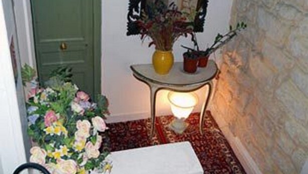 Un pasillo con una puerta verde, una mesita con un florero y plantas en macetas, una lámpara de pie encendida y un arreglo floral.