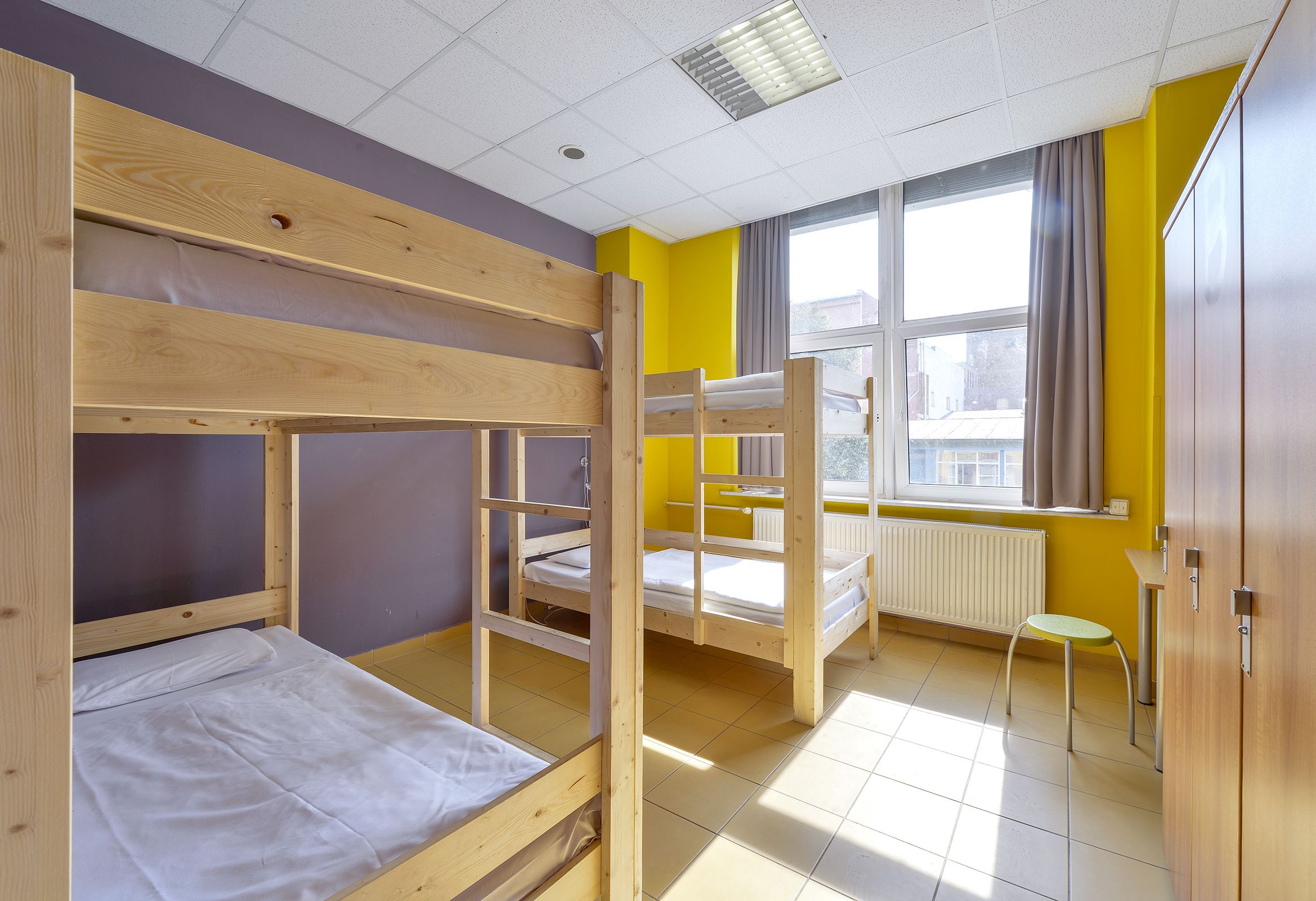 A dormitory room with bunk beds, a window with curtains, a radiator, and a small stool.