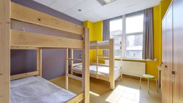 A dormitory room with bunk beds, a window with curtains, a radiator, and a small stool.