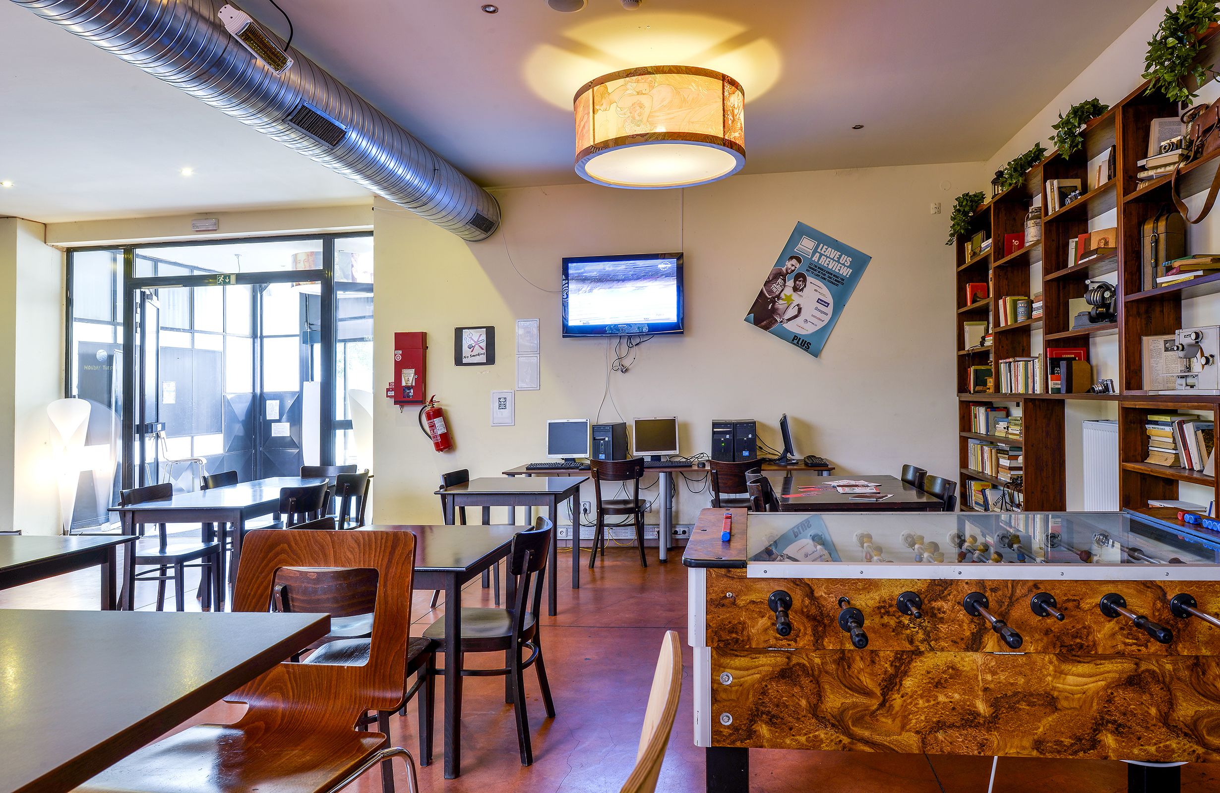Um espaço de escritório moderno com uma janela grande, várias mesas, cadeiras, uma máquina de café e uma estante repleta de livros e objetos decorativos.