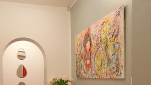 Ein Korridor mit rotem Teppich, einem roten Sofa, einem kleinen weißen Tisch mit einer Froschfigur und einem farbenfrohen, abstrakten Gemälde an der Wand.