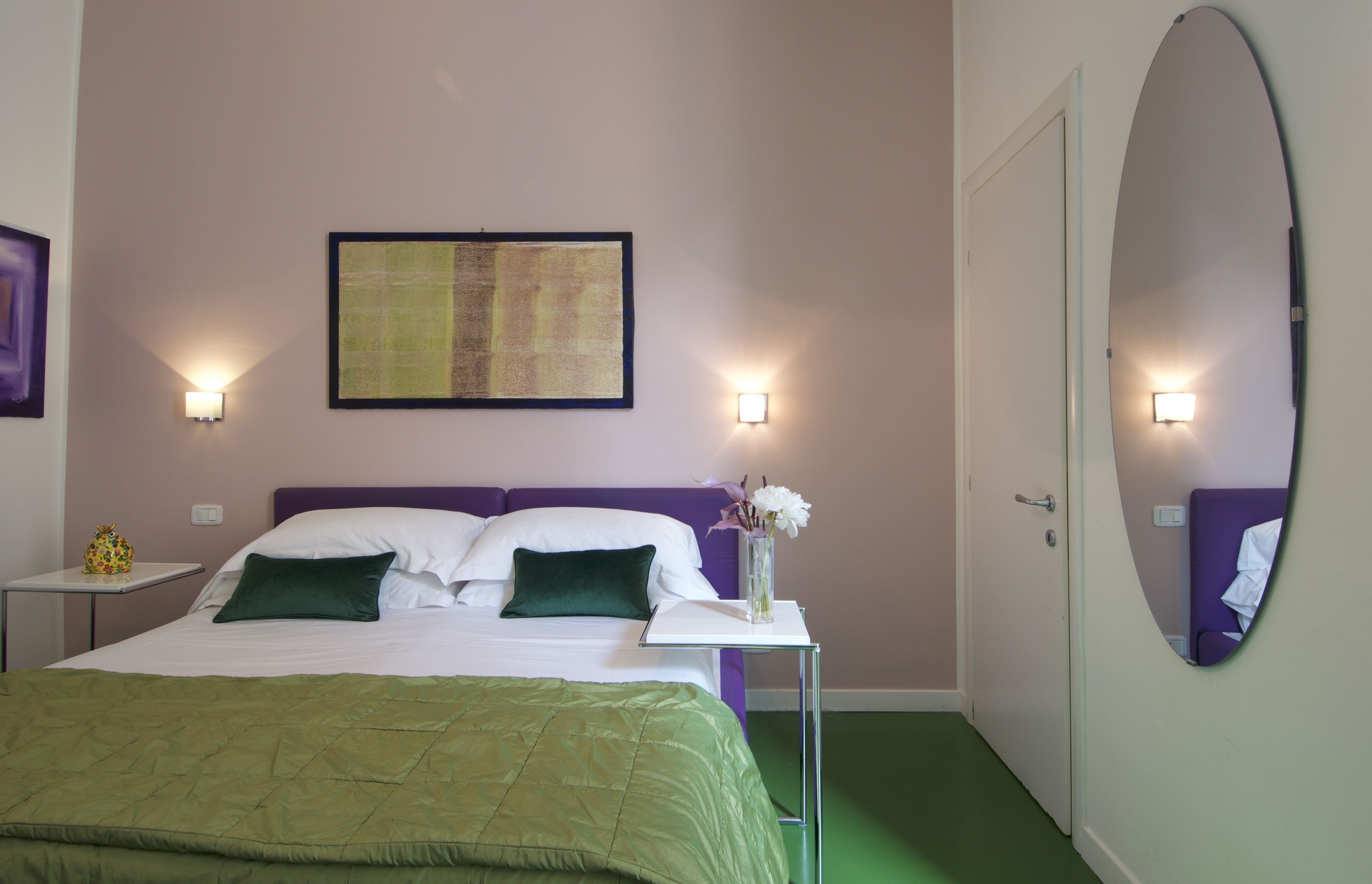 A neatly arranged hotel room with a bed, two pillows, a bedside table with a vase of flowers, and a large mirror on the wall.