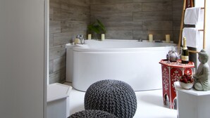 Ein modernes Badezimmer mit einer großen Badewanne, Holzpaneelen, einem kleinen Tisch mit Kerzen und einer Flasche sowie einer dekorativen Buddha-Statue.