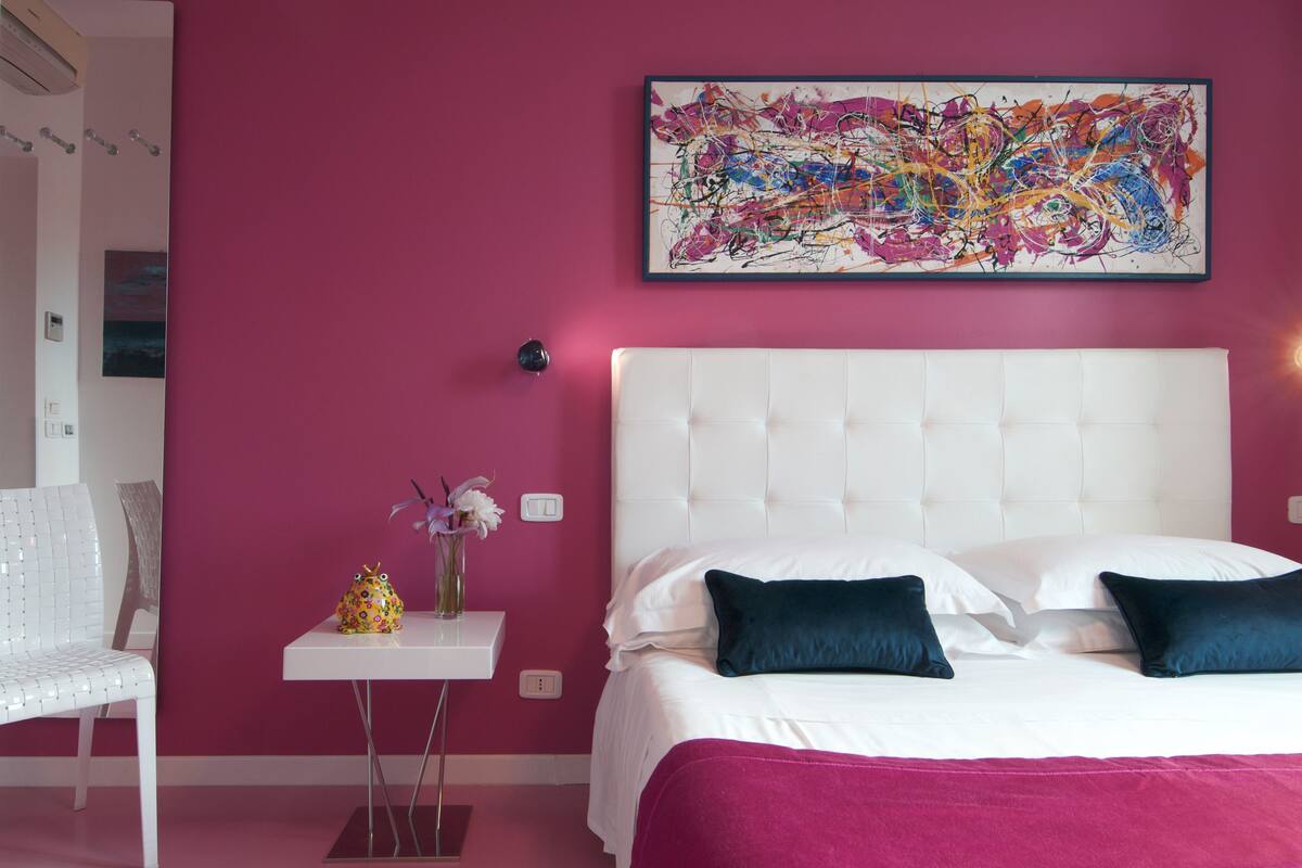 A bedroom with a white bed, pink walls, and a colorful abstract painting.