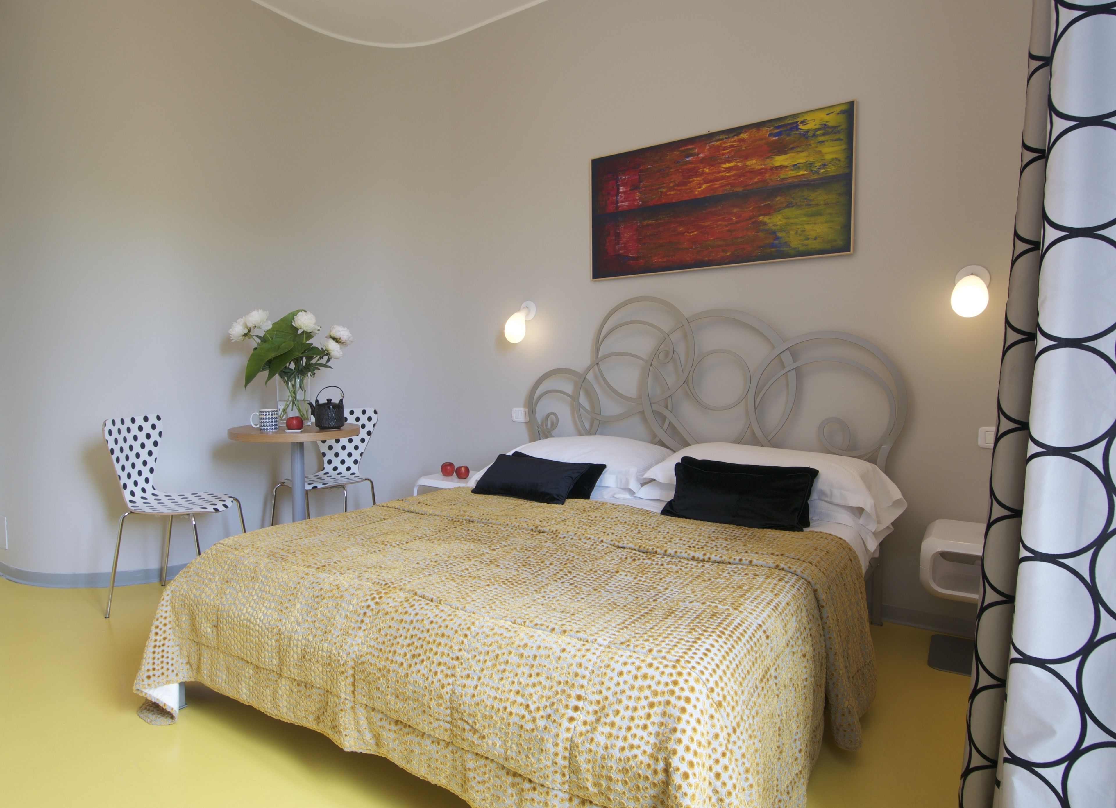 A bedroom with a bed, two chairs, a small table with a vase and apples, and a painting on the wall.