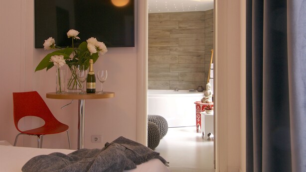 Ein modernes Hotelzimmer mit einem Bett, einem roten Sessel, einem kleinen Tisch mit Blumen, einer Weinflasche und einem Glas. Ein Fernseher ist an der Wand montiert und es bietet sich ein Blick auf ein Badezimmer mit einer Badewanne und einem kleinen Tisch mit einer Statue.