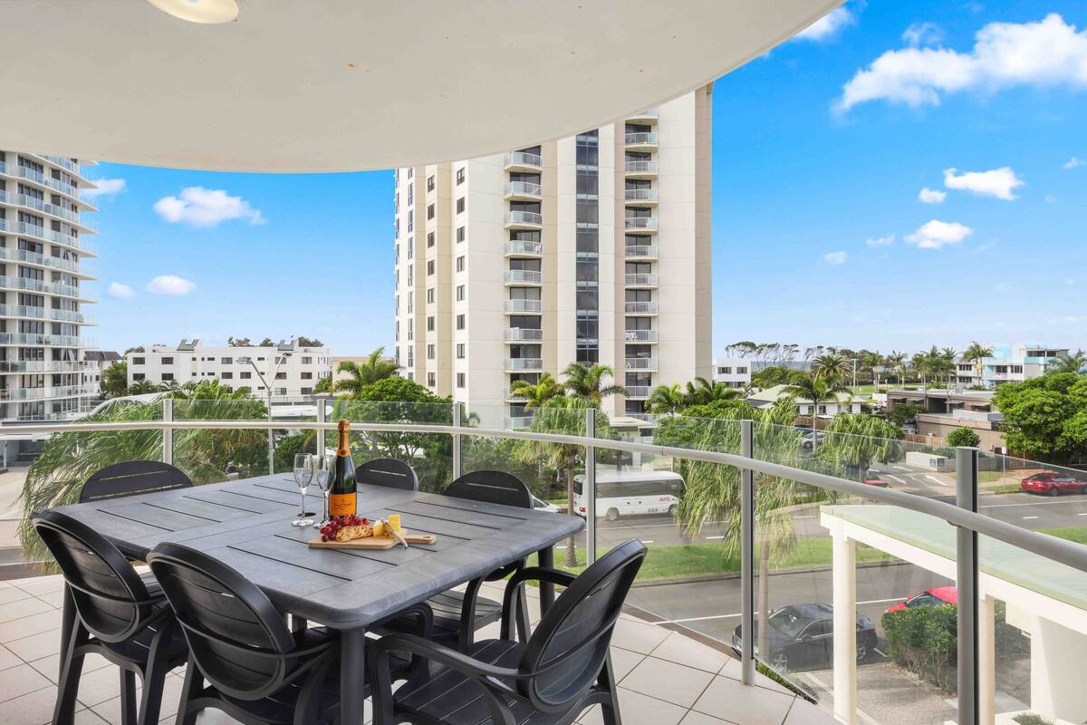 A balcony with a table set for dining, overlooking a cityscape with high-rise buildings and palm trees.