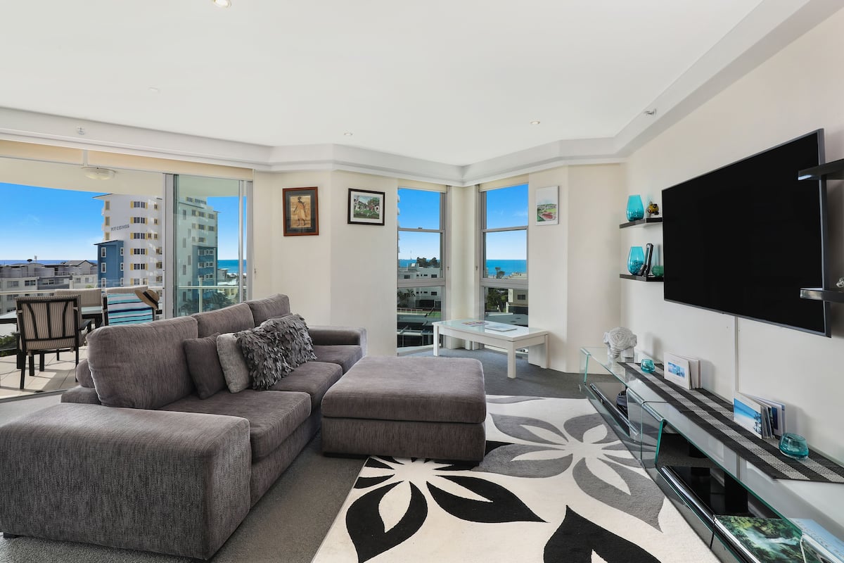 A modern living room with a grey sectional sofa, a glass coffee table, and a large flat-screen TV.