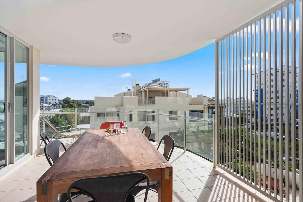 A balcony with a wooden table and chairs, a glass railing, and a view of buildings and greenery.