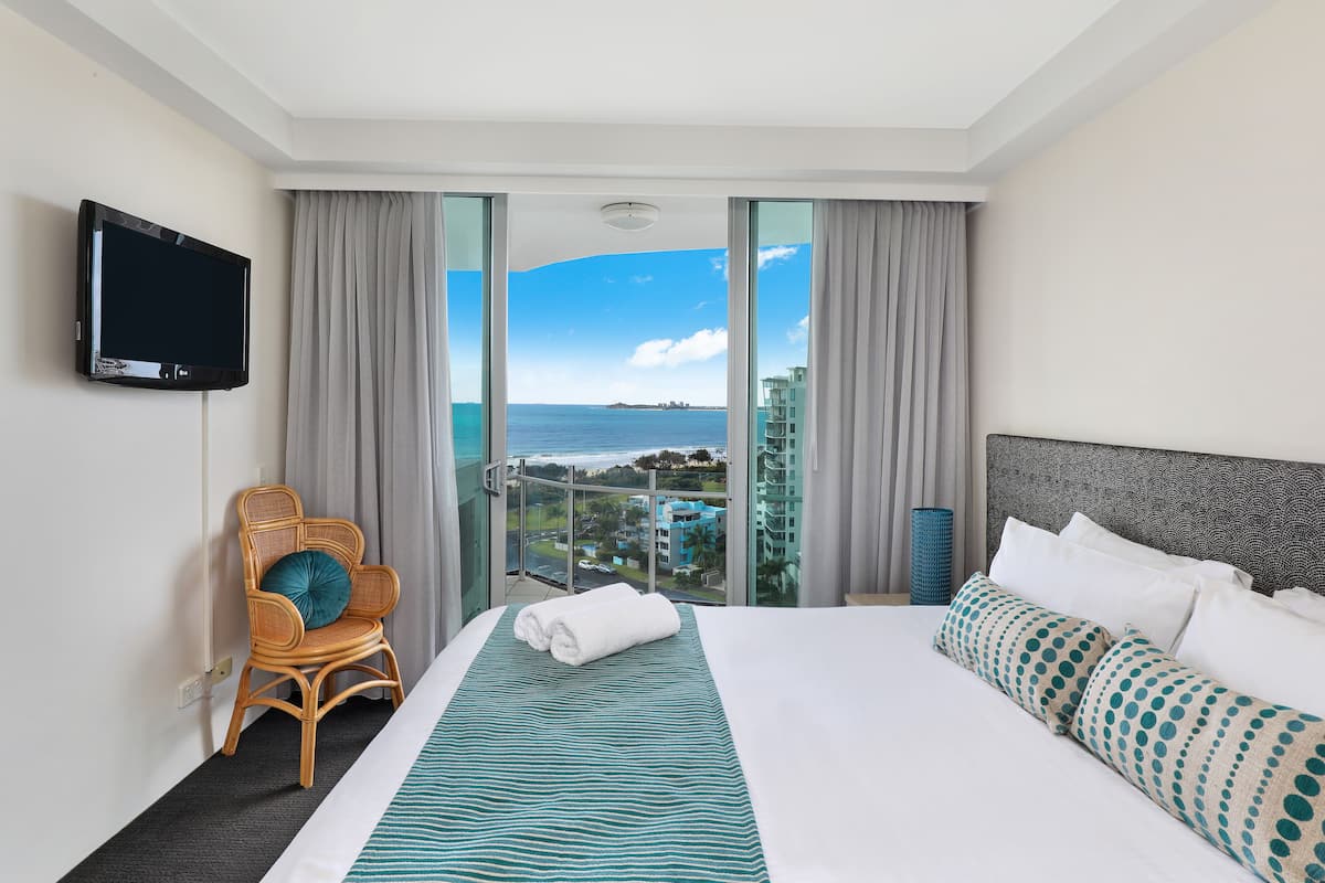 A hotel room with a large bed, a television on the wall, a chair, and a view of the ocean.