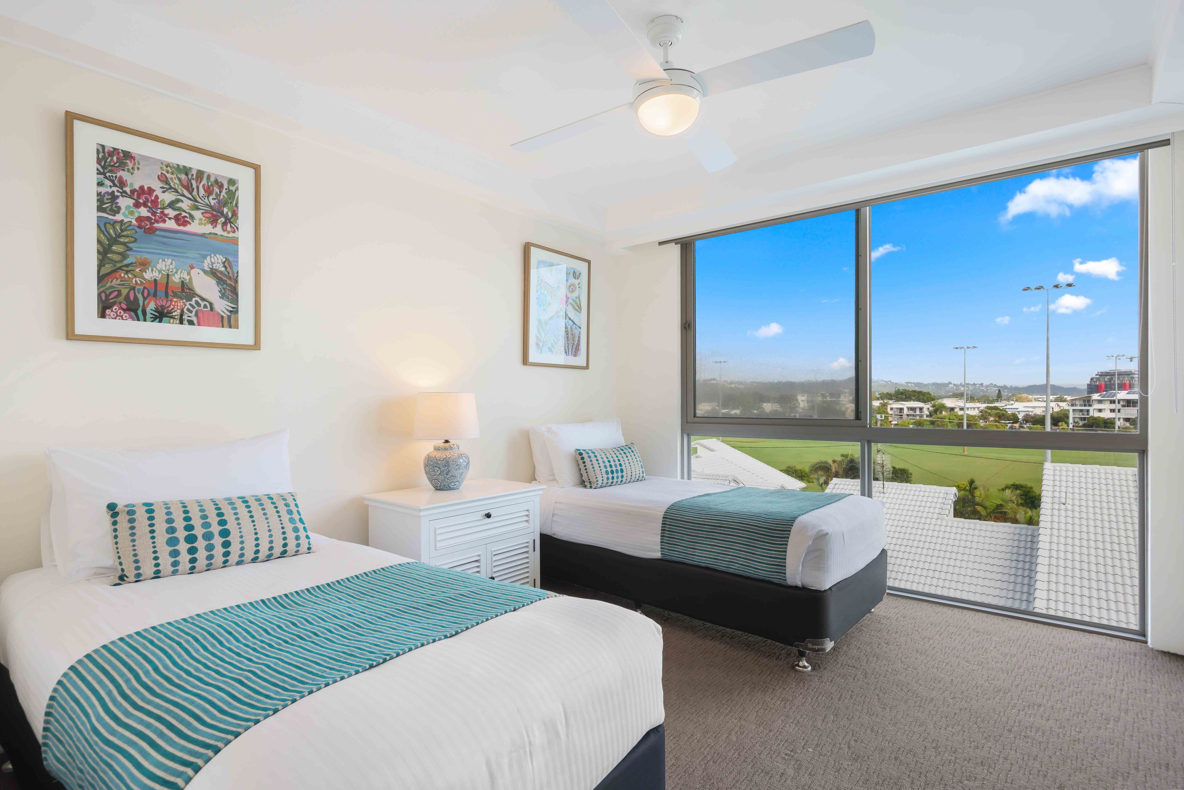 A hotel room with two beds, a ceiling fan, a large window with a view of the city, and artwork on the walls.