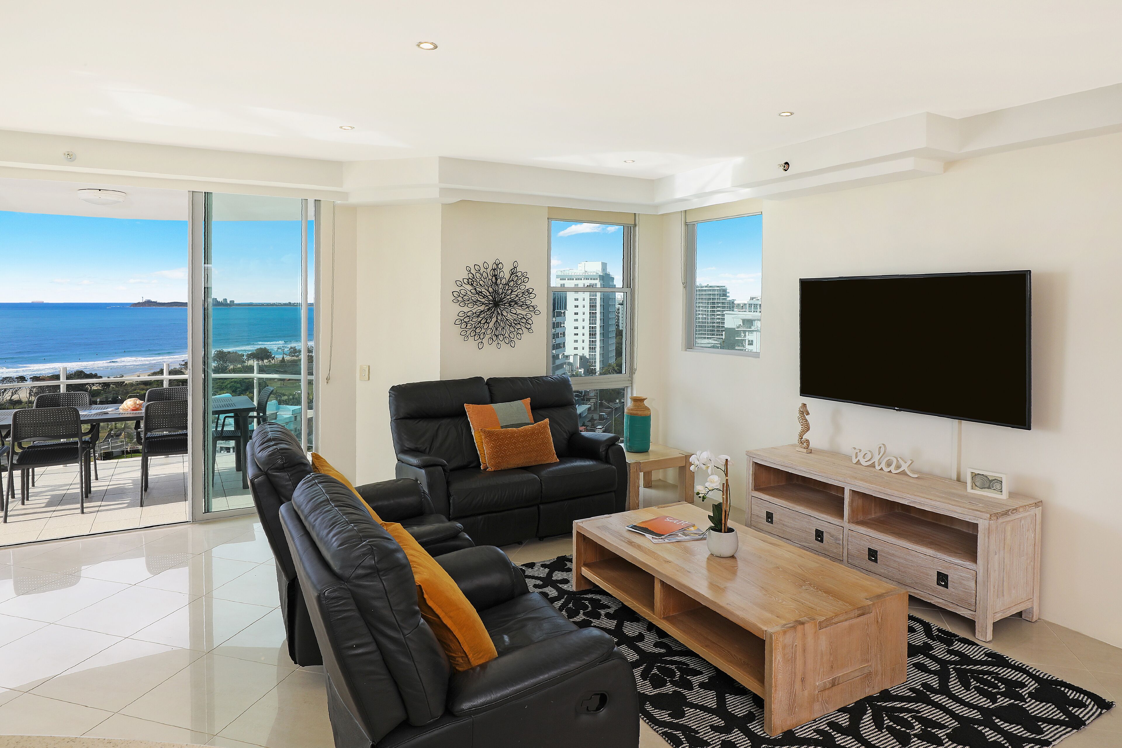A modern living room with a large TV, black leather sofa, wooden coffee table, and a view of the ocean.
