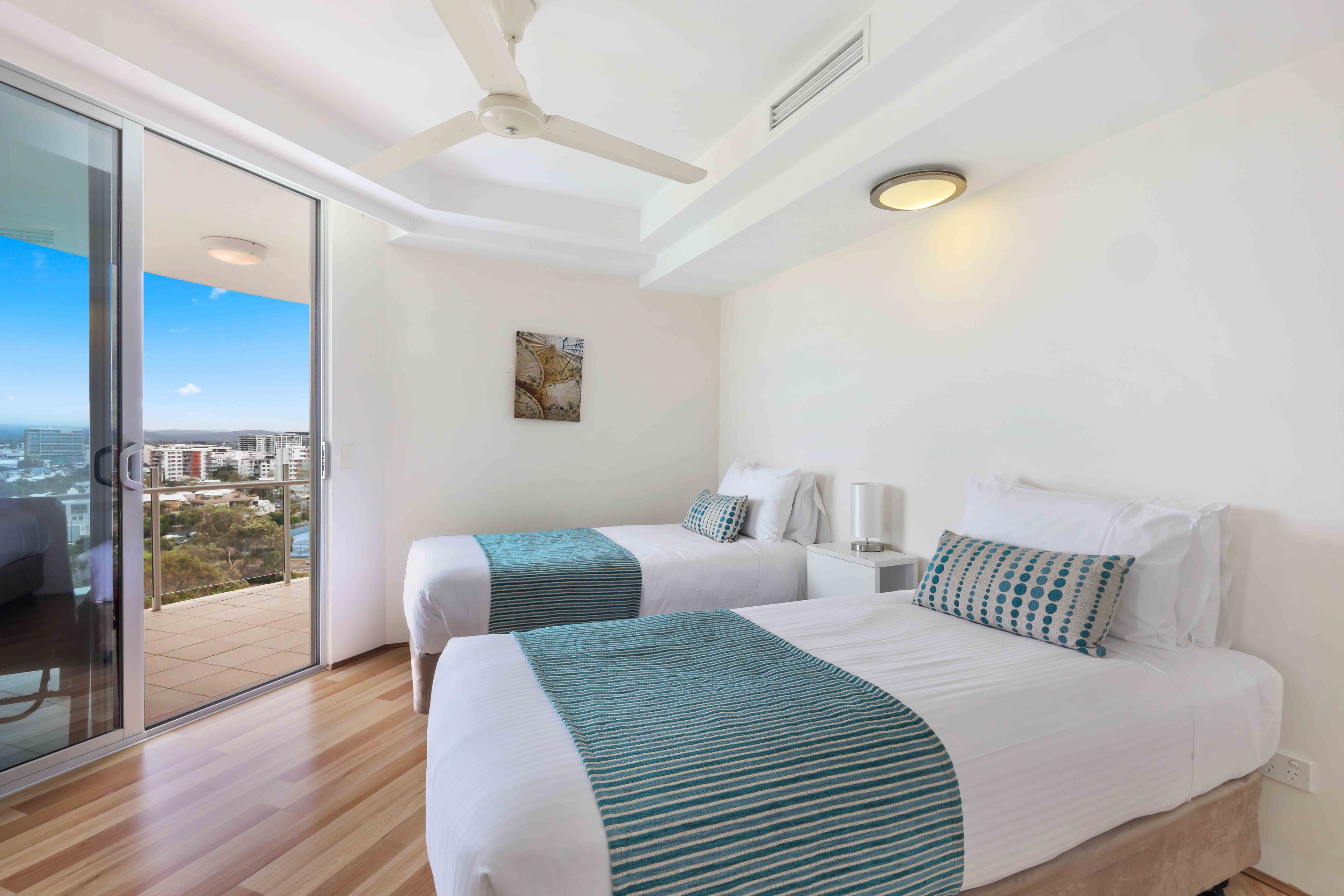 A modern hotel room with two beds, a balcony with a city view, a ceiling fan, and a painting on the wall.