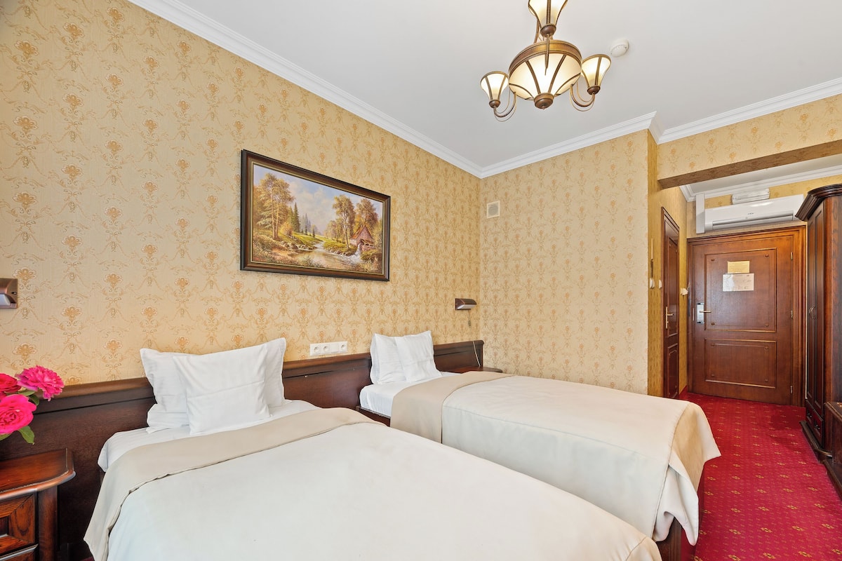 A hotel room with two beds, a painting on the wall, a chandelier, and a red carpet.