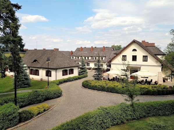 Un edificio de estilo europeo tradicional con un camino adoquinado que conduce a un patio, rodeado de setos y árboles bien cuidados.