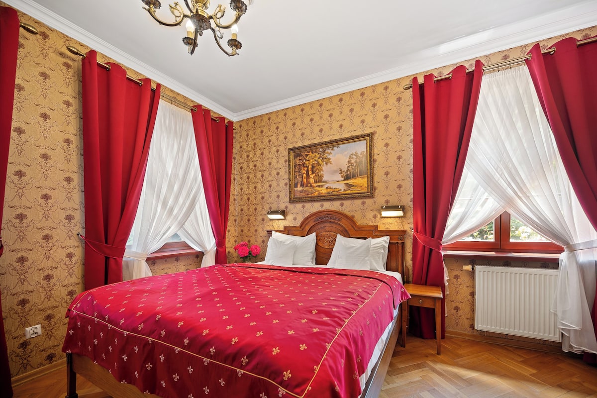 A bedroom with a large bed, red curtains, a wooden headboard, a nightstand, a bedside table, a radiator, and a painting on the wall.