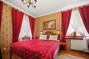 Un dormitorio con una cama grande, cortinas rojas, un cabecero de madera, una mesita de noche y un radiador. Hay un cuadro en la pared.