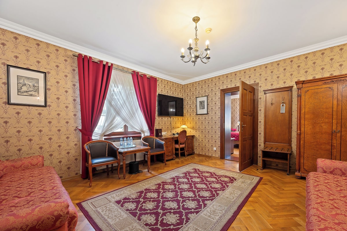 A room with a wooden floor, a bed with a red bedspread, a desk with a chair, a wooden wardrobe, and a chandelier.