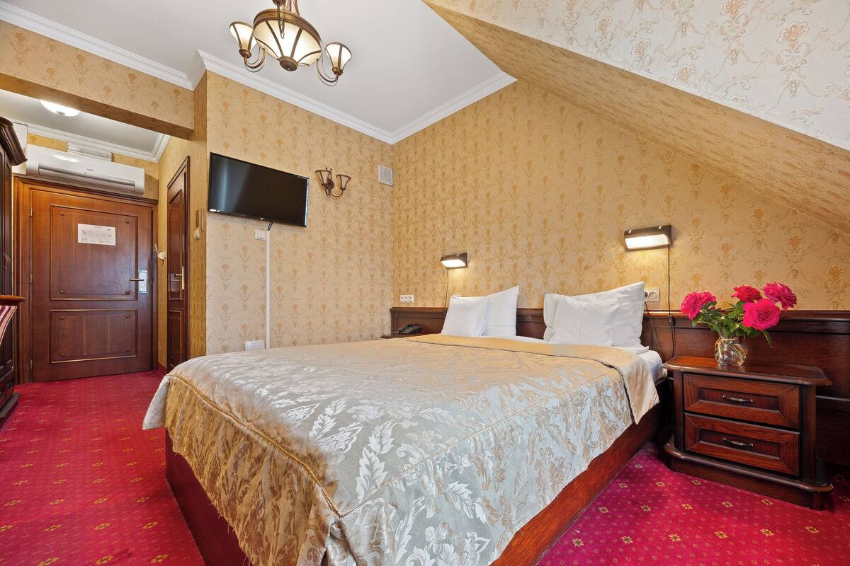 A hotel room with a large bed, a wooden nightstand, a TV mounted on the wall, and a red carpeted floor.
