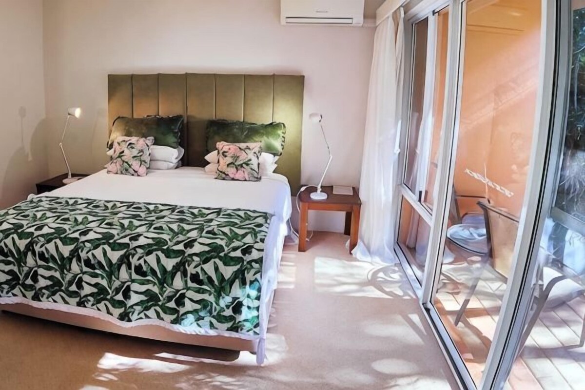 Un dormitorio con cama, almohadas, una mesita de noche, una lámpara y un ventanal corredizo que da a un balcón.