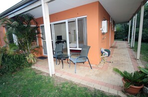 Un patio cubierto con una mesa y sillas, un ventanal corredera que da a una habitación, y un jardín con plantas en macetas.